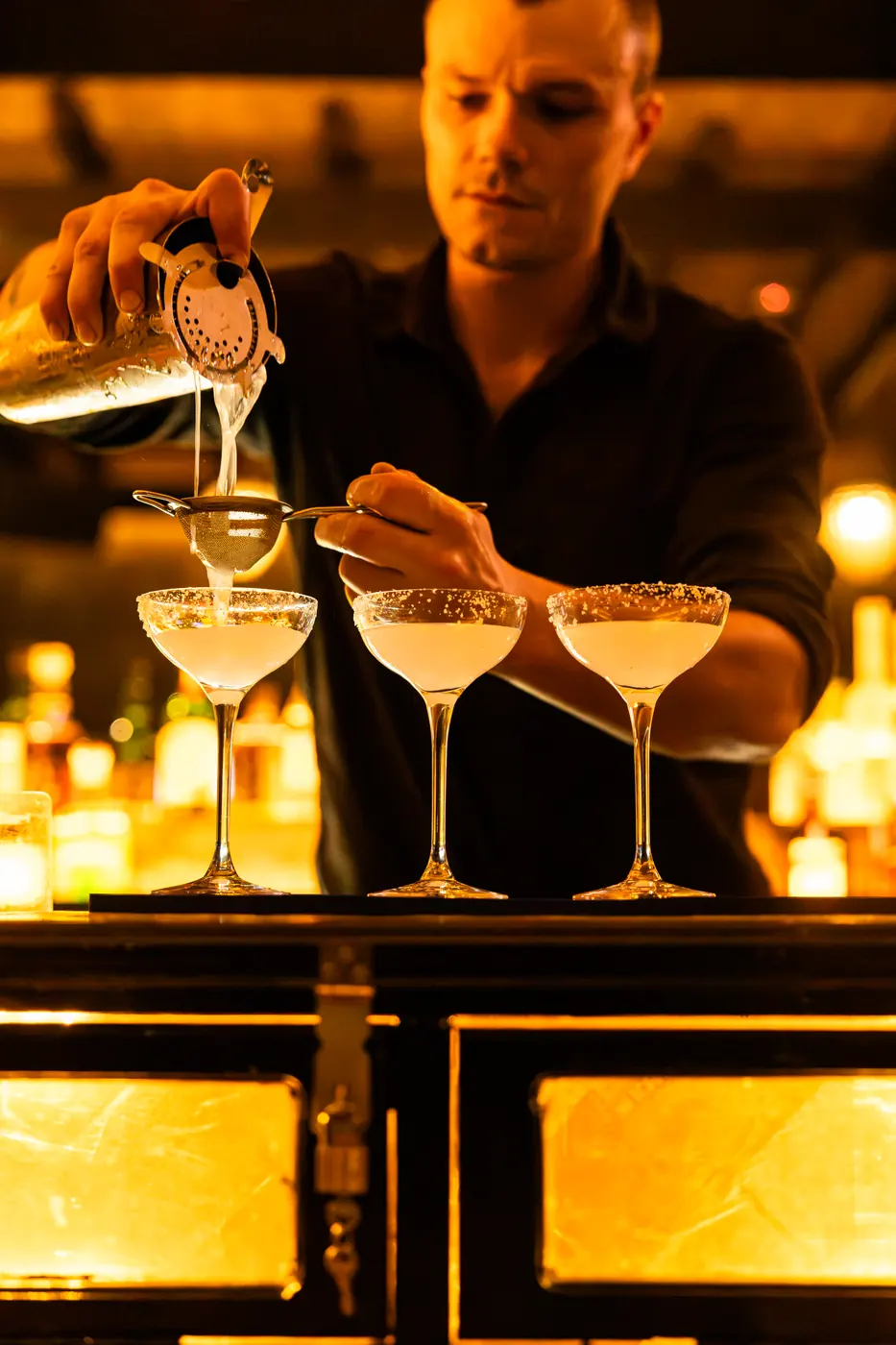 A bartender straining cocktails into three coupes at the Soi Fire bar
